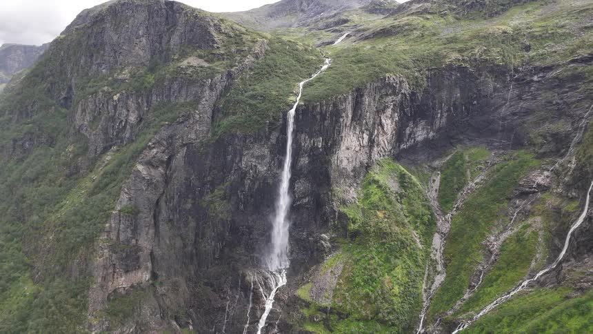 A powerful waterfall tumbles down a sheer rock cliff in a lush, green mountain valley in Norway as the camera slowly zooms out