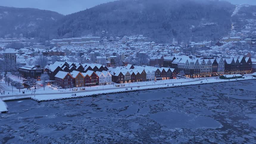 A static wide shot of the historic Bryggen wharf in Bergen, Norway, covered in snow during a winter twilight, with ice floating in the harbor