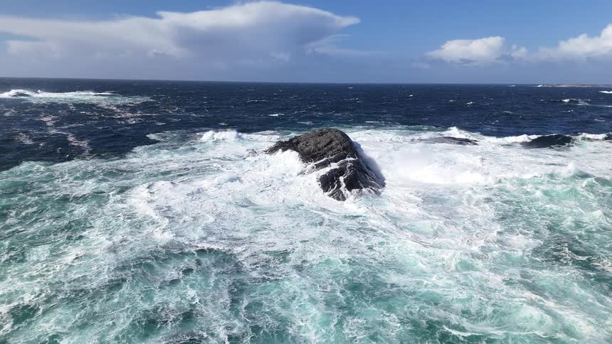 A mesmerizing slow motion aerial shot captures the raw power of the ocean as turquoise waves crash over a solitary rock
