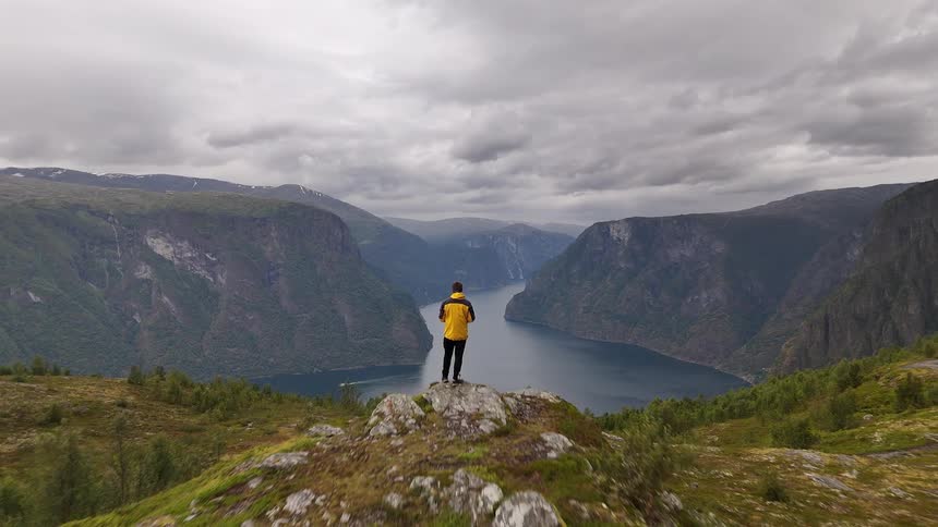 Dramatic aerial reveal of a man in a yellow jacket standing on a rocky outcrop, admiring the vast and beautiful Norwegian fjord