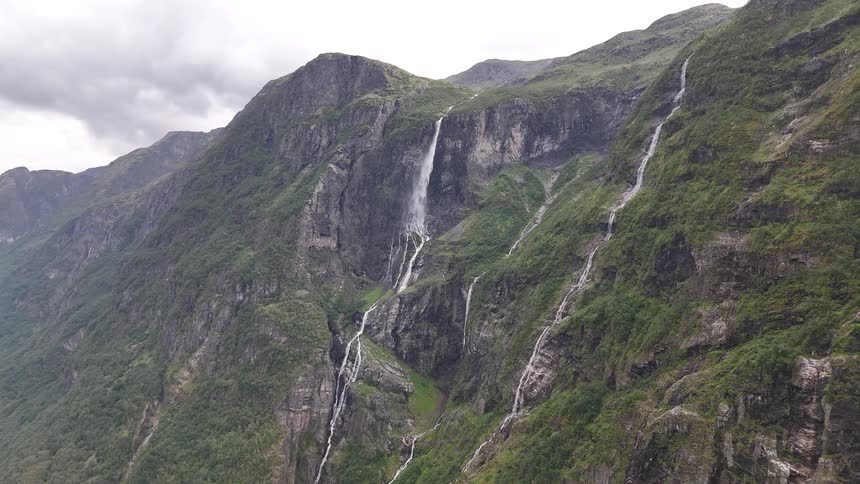 Multiple powerful waterfalls cascade down a steep, green, and rocky mountainside under an overcast sky