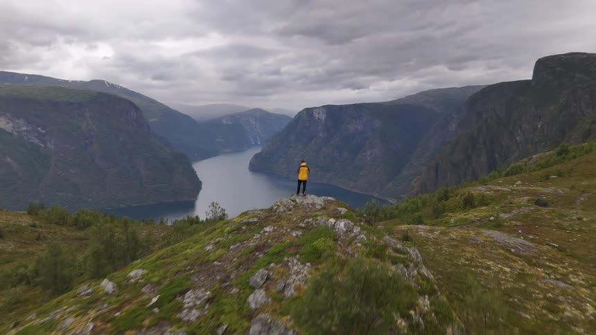 An aerial view pushes past a hiker in a yellow jacket standing on a rocky outcrop to reveal the stunning Aurlandsfjord in Norway on a cloudy day