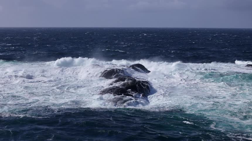 Powerful white ocean waves crash over a dark rock in the deep blue sea in dramatic slow motion