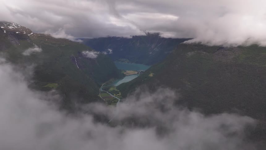Drone flies through dense fog to reveal a stunning high-altitude view of a vast green valley, river, and lake surrounded by mountains