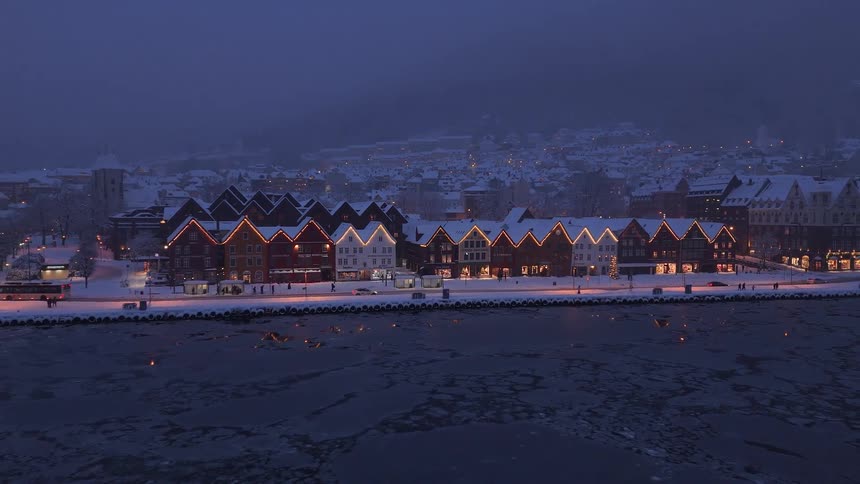 A charming coastal village glows with festive lights on a snowy winter evening as ice floes drift in the harbor