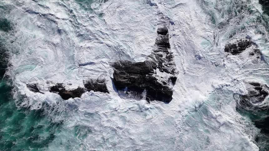 Hypnotic top-down aerial view of a powerful ocean vortex as waves crash over dark rocks in slow motion