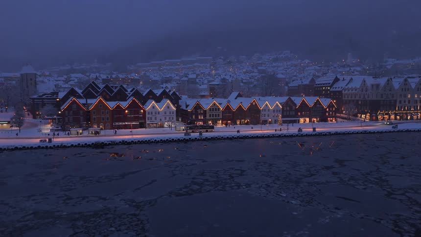 The iconic Bryggen waterfront in Bergen, Norway, is covered in snow and adorned with festive lights on a cold winter evening as ice floats in the harbor