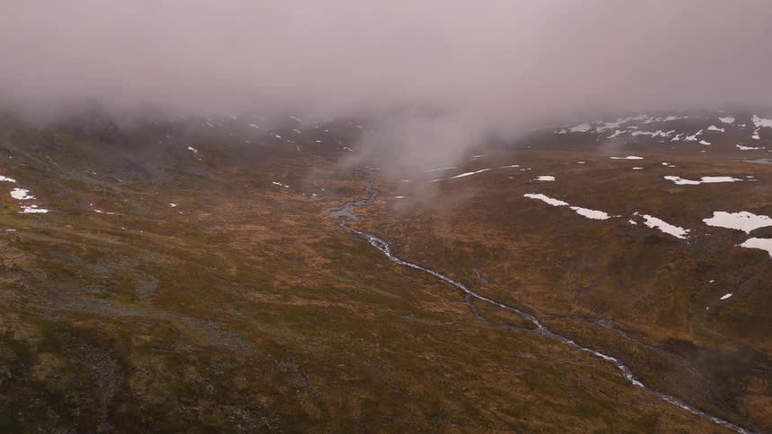 A stationary aerial view of a misty, desolate mountain valley with a winding stream and patches of snow