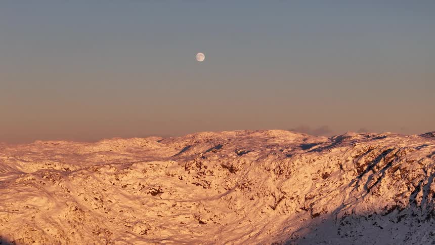 The full moon hangs in the clear sky over a vast, snow-covered mountain range glowing in the warm light of sunrise
