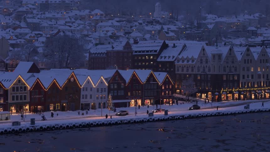 The historic Bryggen wharf in Bergen, Norway, glows with festive lights on a snowy winter evening as people and cars move along the harbor