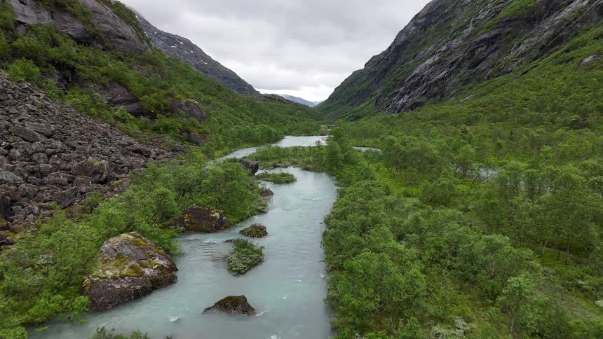 A slow aerial shot flies over a turquoise glacial river winding through a lush green valley between steep mountains
