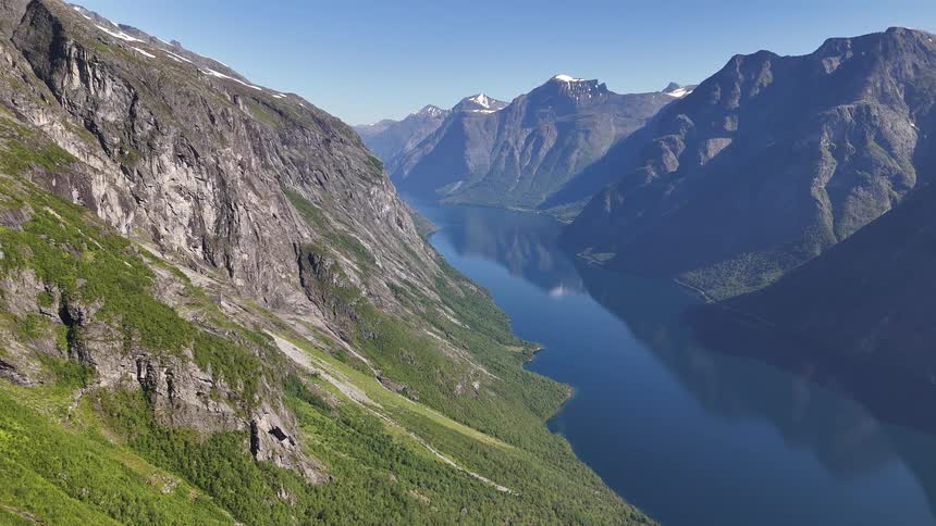 A breathtaking high-angle aerial view slowly reveals the immense scale of a tranquil fjord nestled between towering, majestic mountains in Norway