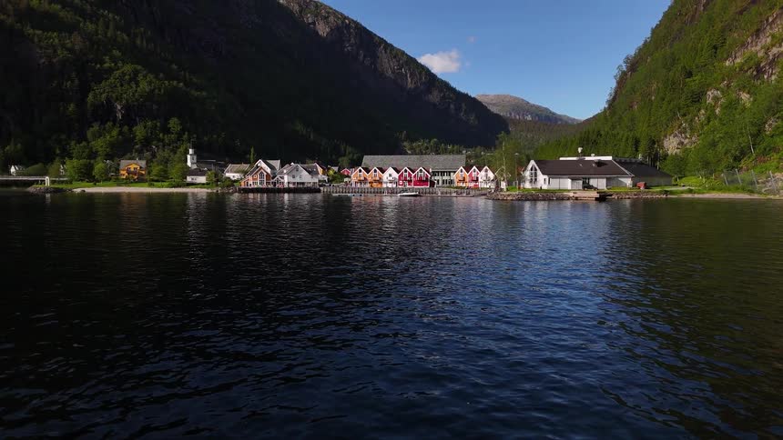 An aerial drone shot glides over the calm waters towards a picturesque village with vibrant houses on the shore of a Norwegian fjord