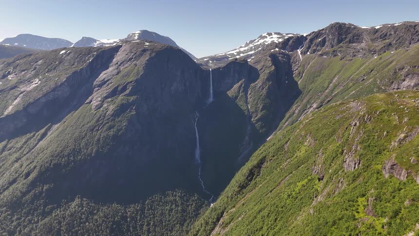 A breathtaking aerial view captures a powerful waterfall cascading down a rugged, snow-capped mountain into a lush green valley on a clear sunny day