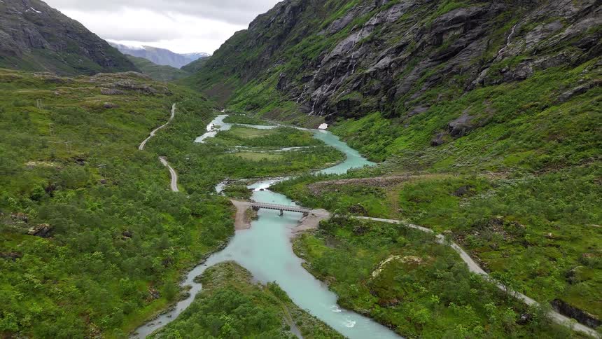 A stunning aerial pullback shot reveals a turquoise glacial river winding through a lush green valley between majestic mountains