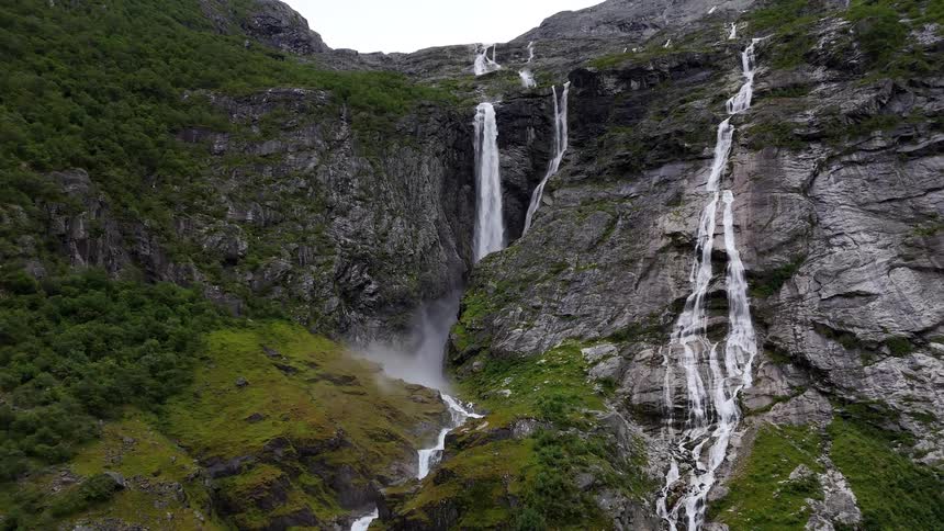 A slow aerial pullback reveals multiple powerful waterfalls cascading down a rugged, moss-covered mountain in Norway