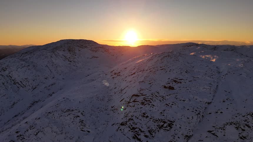 A majestic aerial view shows the sun rising over a vast, snow-covered mountain range, casting a golden light on the peaks