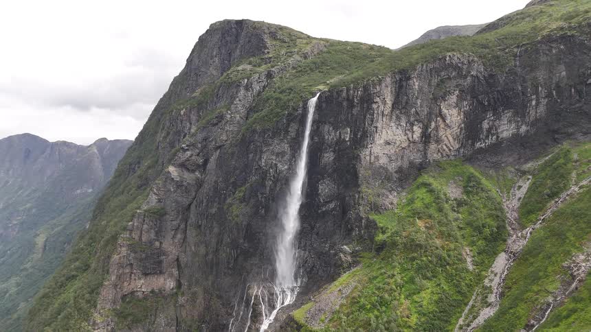 An epic wide shot captures a powerful waterfall flowing down a massive, green mountain cliff into a fjord below