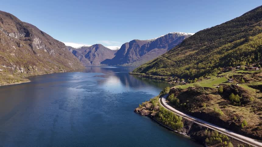 A stunning static aerial view of a Norwegian fjord, revealing deep blue waters, green mountainsides, and a winding coastal road under a clear sky
