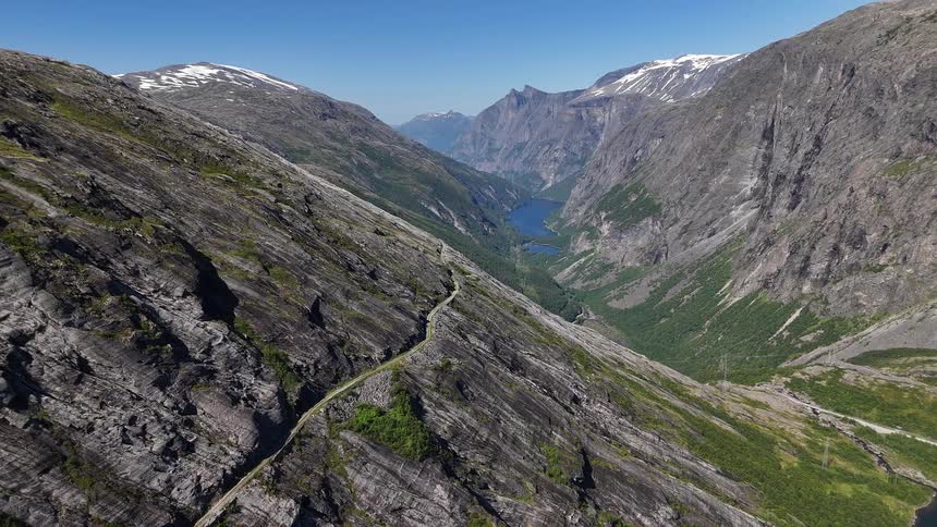 A stunning aerial view of a winding road through a majestic, rocky mountain valley in Norway with a lake in the distance