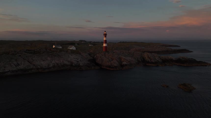 An aerial view of a picturesque red and white lighthouse shining its light over the ocean from a rocky coast during a beautiful sunset