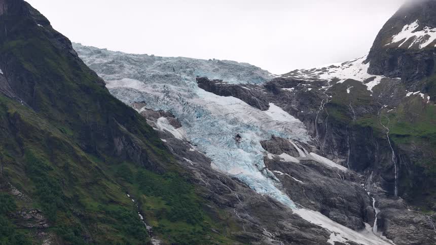 A static wide shot captures the immense Boyabreen glacier nestled between lush green mountains in Norway