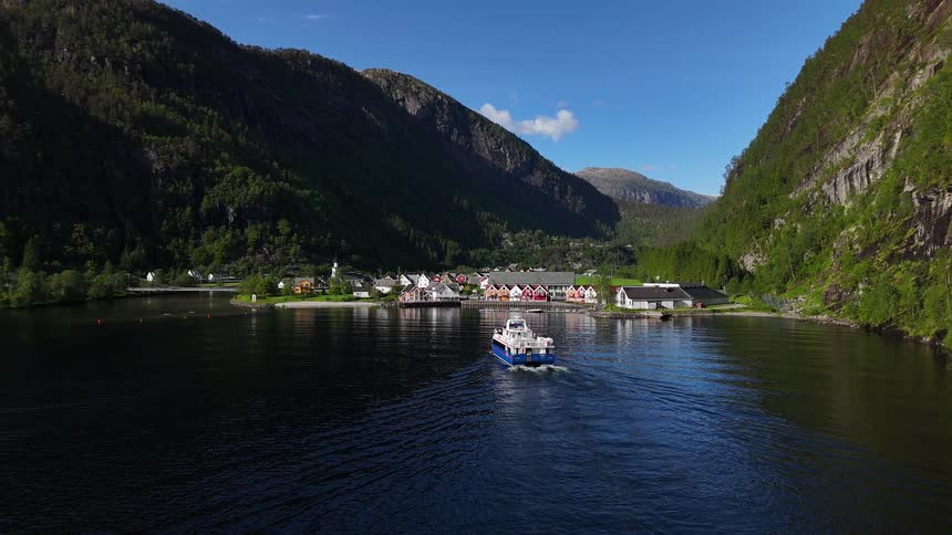 A passenger ferry sails across a serene fjord towards a charming coastal village nestled between steep, green mountains on a sunny day
