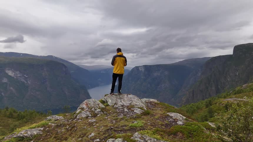 A man in a yellow jacket stands on a rocky mountain overlook, taking in the epic view of a Norwegian fjord under a cloudy sky