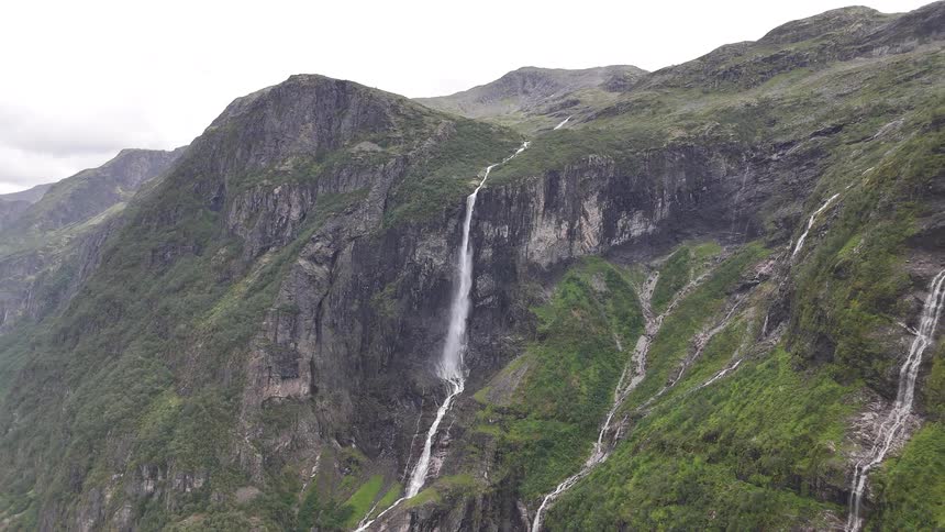 Majestic waterfalls cascade down the steep, rocky cliffs of a lush green mountain under a cloudy sky