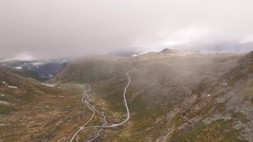 An aerial view rises through thick fog to reveal a vast mountain landscape with a winding road and river