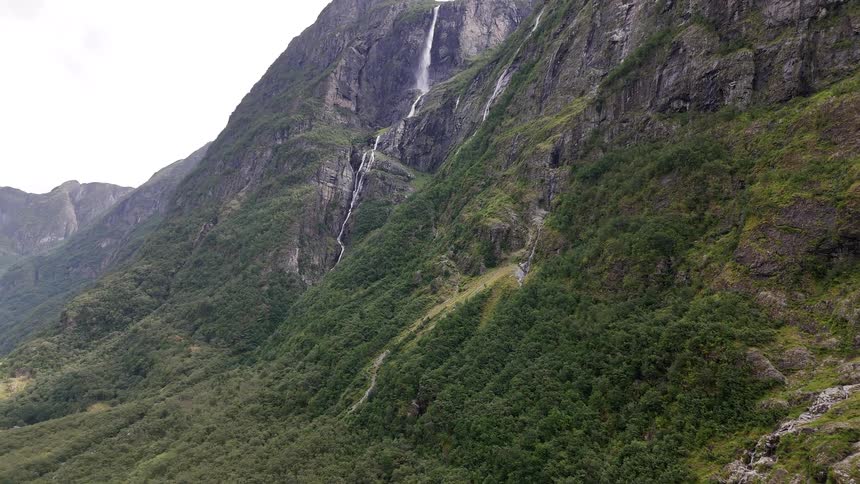 A slow aerial tilt-up reveals several majestic waterfalls cascading down a steep, verdant mountain into a lush valley below