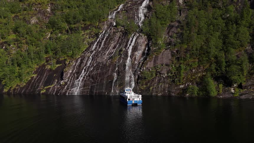 A tourist catamaran boat floats peacefully near a stunning waterfall cascading down a steep, forested cliff in a Norwegian fjord