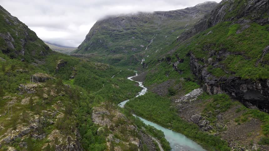 Epic aerial footage shows a winding turquoise river flowing through a dramatic, lush green mountain valley in Norway