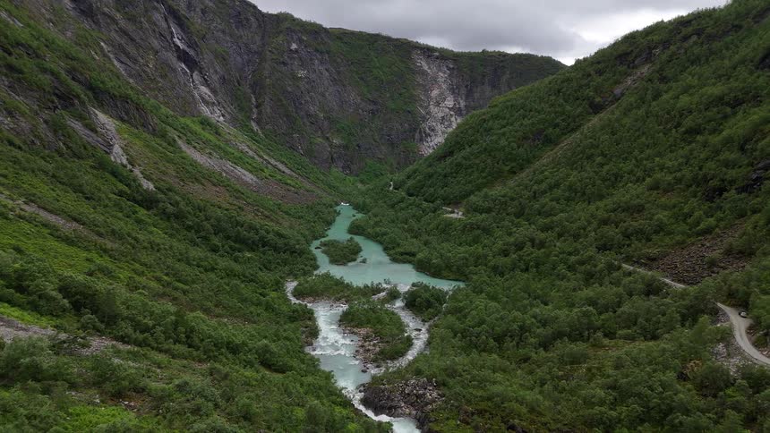 An epic aerial view slowly reveals a turquoise glacial river flowing through a deep, lush green valley in the mountains of Norway