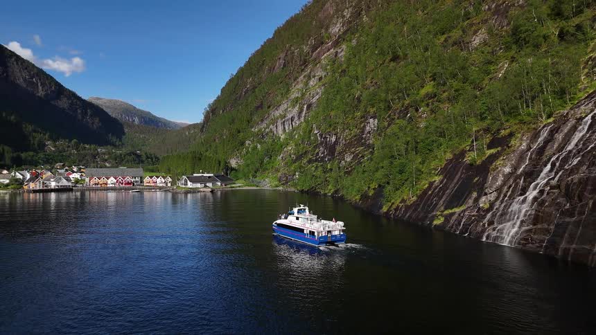 A scenic aerial view follows a passenger ferry sailing past a magnificent waterfall and towards a quaint village on a Norwegian fjord