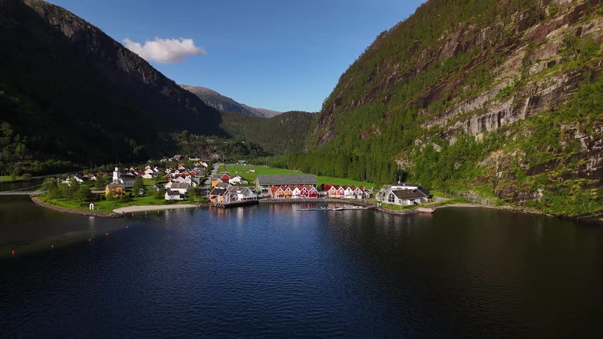 An idyllic Norwegian village with colorful houses nestled on the serene waters of a fjord between towering green mountains on a bright sunny day