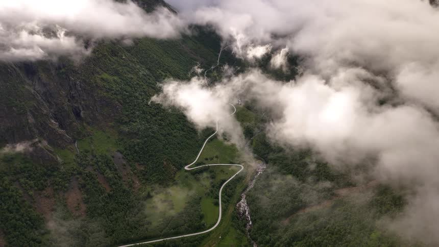 An epic aerial shot peers through the clouds at a serpentine road winding through a majestic green valley