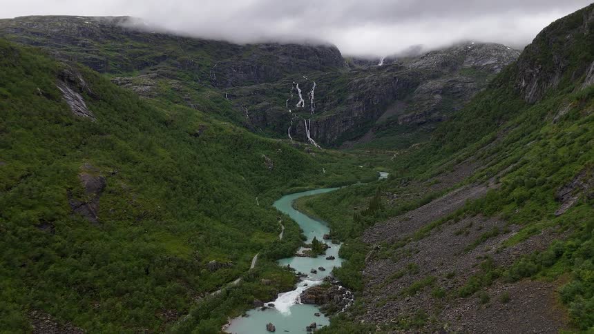 A majestic aerial shot captures a turquoise glacial river winding through a lush green valley in Norway, with waterfalls cascading down the misty mountains