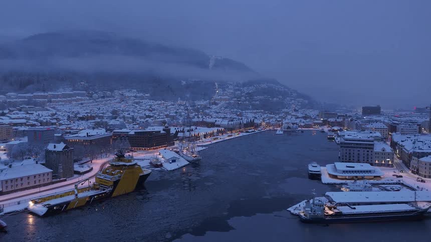 A serene aerial view reveals a snow-covered coastal city and its partially frozen harbor at twilight during winter