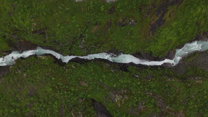 An aerial top-down view of a powerful glacial river carving a path through a lush green canyon, fed by a cascading waterfall