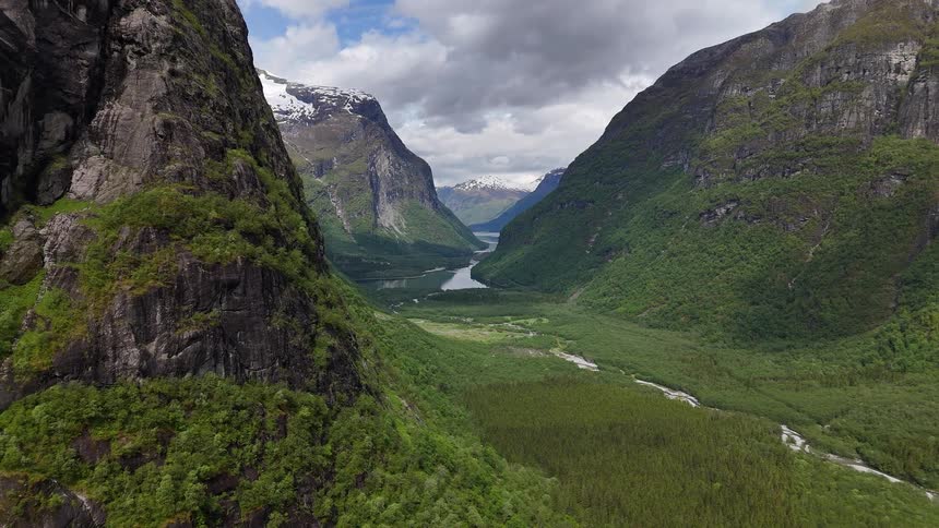 A majestic aerial view captures a lush green valley with a winding river nestled between towering, snow-capped mountains in Norway
