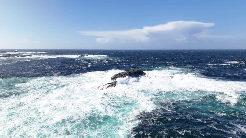 Powerful ocean waves crash in slow motion over a rugged rock formation on a bright sunny day