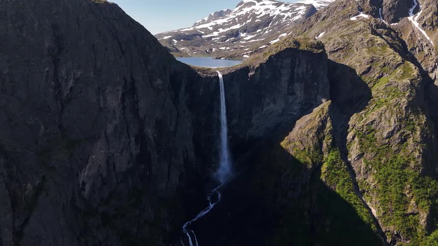 An epic aerial view reveals a stunning waterfall cascading from a high mountain lake into a rocky valley