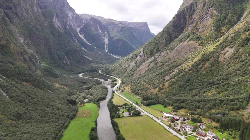 A majestic aerial view captures a river winding through a lush green valley with a small village, surrounded by towering mountains