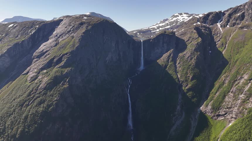 A breathtaking static aerial view of a powerful waterfall cascading down a vast, lush green mountain range with snow-capped peaks