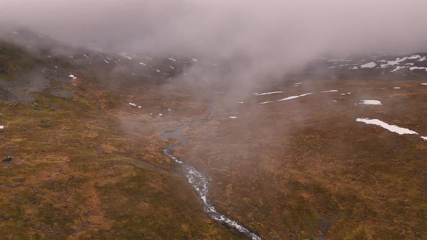 Thick fog rolls through a desolate mountain valley where a river flows through the barren landscape with patches of lingering snow