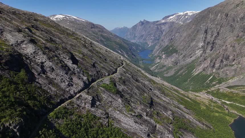 A breathtaking aerial shot slowly reveals an epic and vast mountain valley landscape with a winding road and distant lakes under a clear blue sky