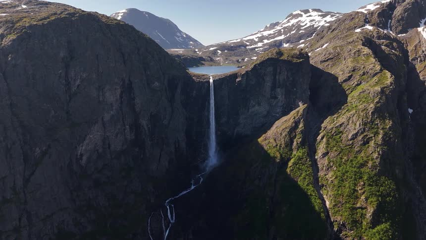 A majestic aerial view shows a powerful waterfall cascading from a high mountain lake down a steep cliff into a winding river below