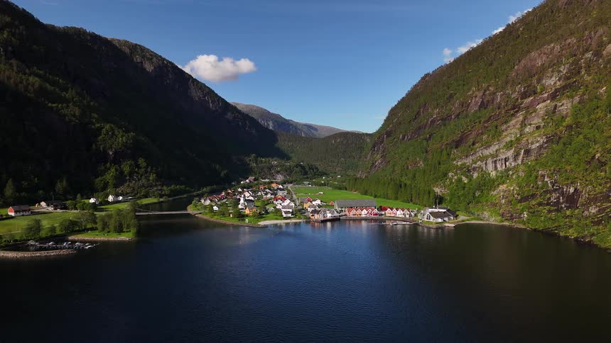 A stunning static aerial shot captures the serene beauty of a small Norwegian village on the banks of a fjord, flanked by imposing green mountains
