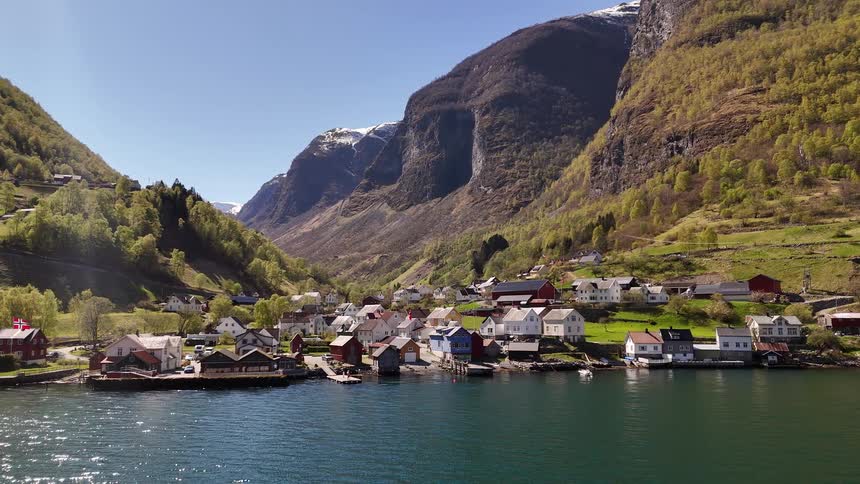 A tranquil Norwegian village sits on the shore of a stunning fjord, nestled between majestic mountains under a clear blue sky